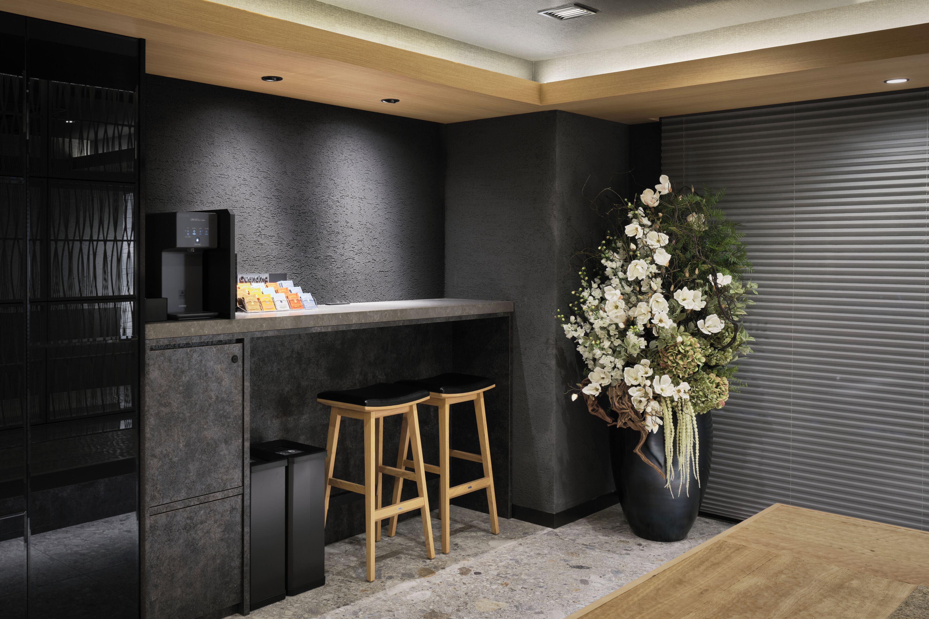 A modern kitchen with dark walls, a black countertop, two wooden stools, a coffee machine, and a large potted plant with white flowers.