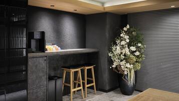 A modern kitchen with dark walls, a black countertop, two wooden stools, a coffee machine, and a large potted plant with white flowers.