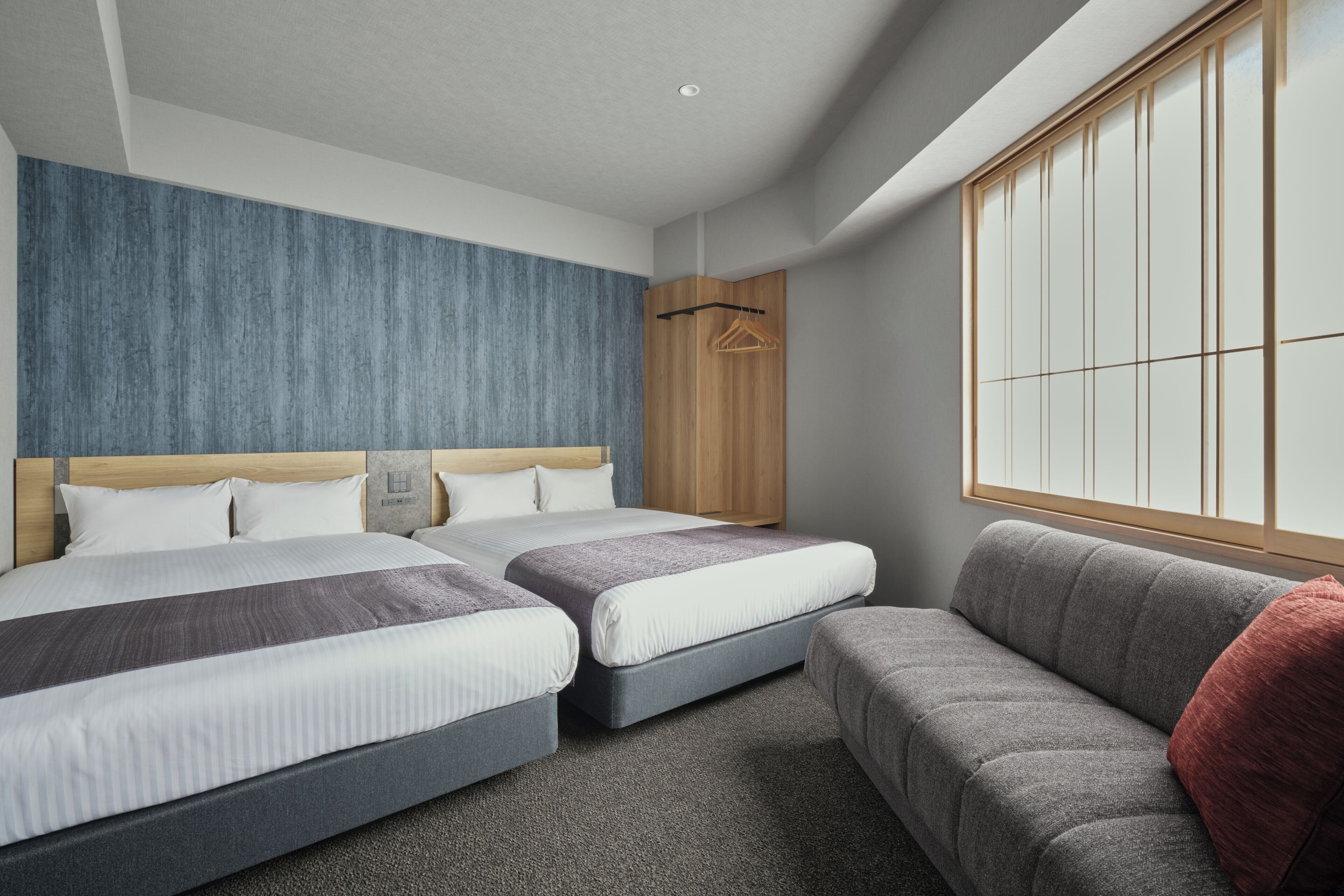 A hotel room with two beds, a sofa, a wooden wardrobe, and a large window.