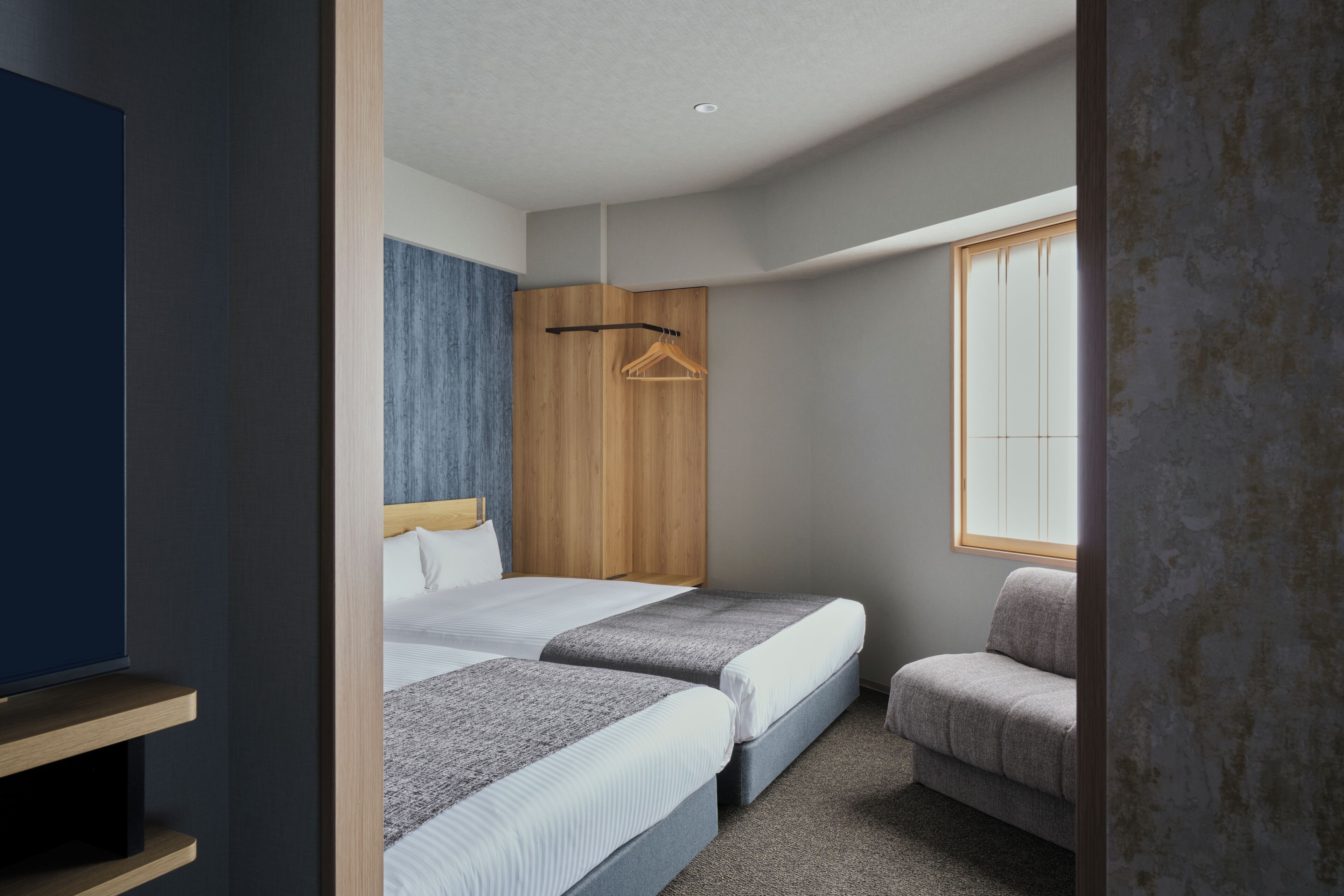 A modern hotel room with two beds, a wooden wardrobe, a seating area, and a television.