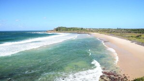 Une plage côtière aux eaux limpides, bordée d’un rivage sablonneux, avec une colline verdoyante à l’horizon.