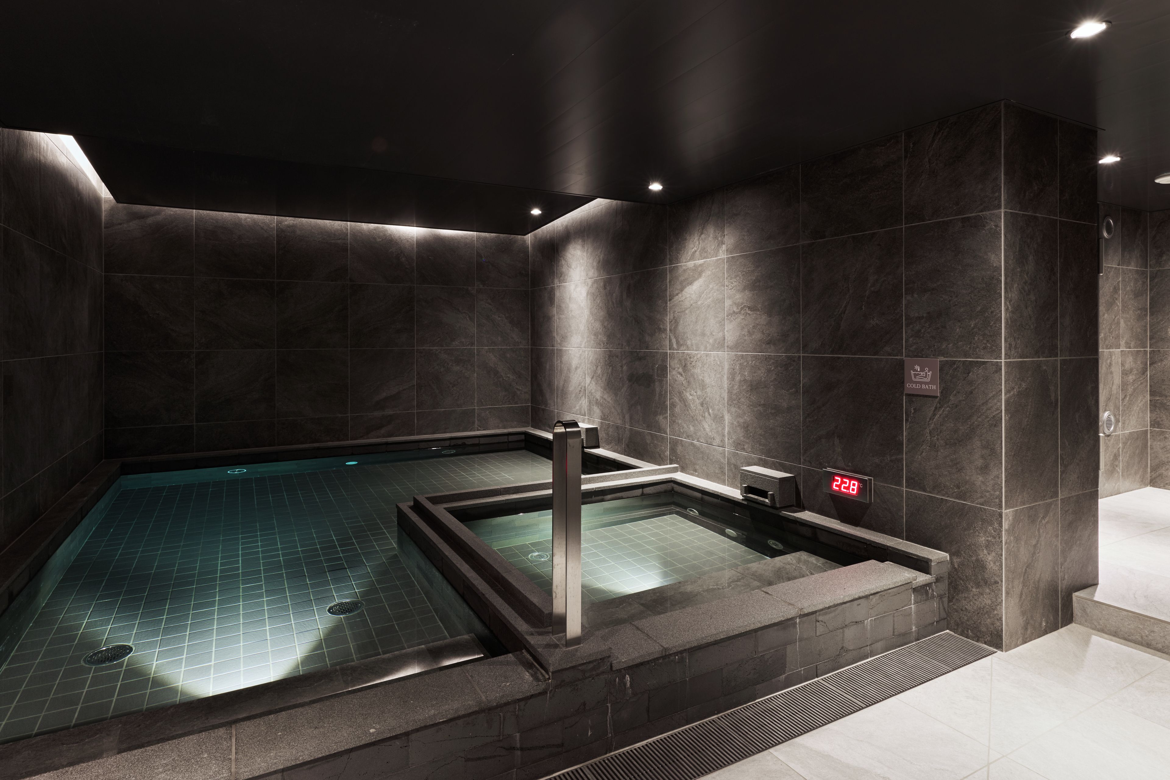 A modern spa area with a rectangular hot tub, dark stone walls, and a digital thermometer indicating 22.8°C.