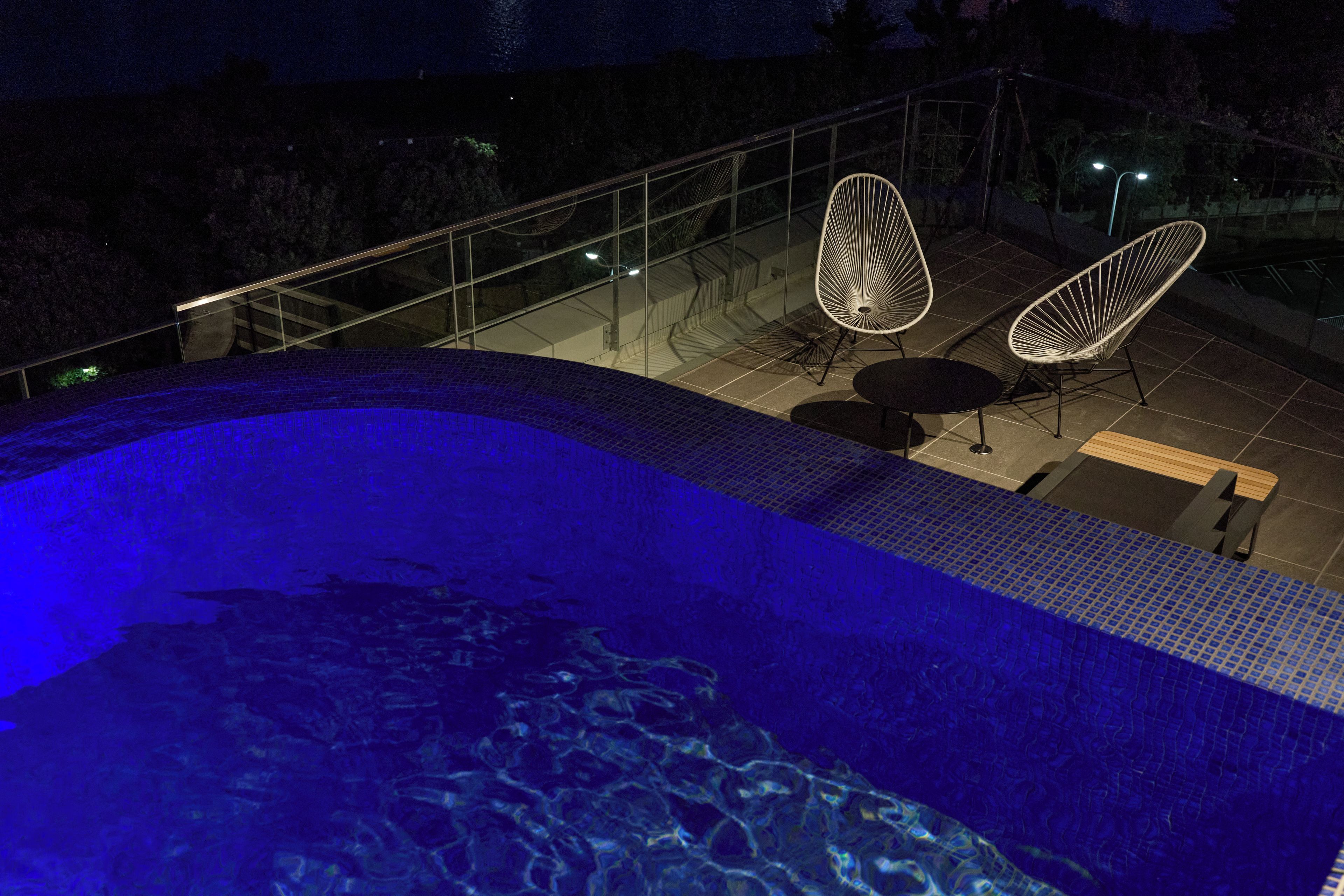 A swimming pool with a glass balustrade, two lounge chairs, and a small table.