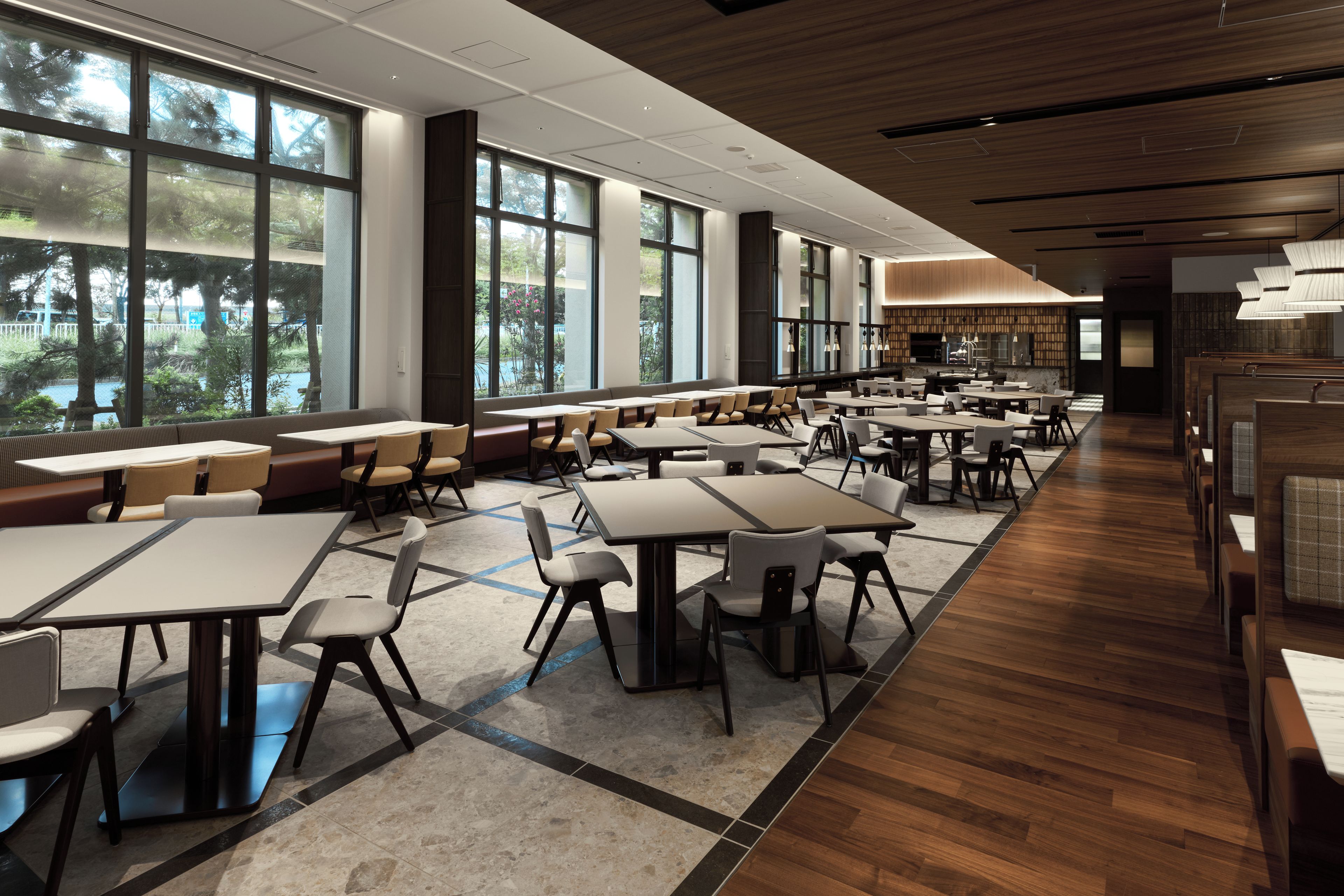A spacious indoor dining area with multiple tables and chairs, large windows offering views of greenery, and a modern ceiling design.