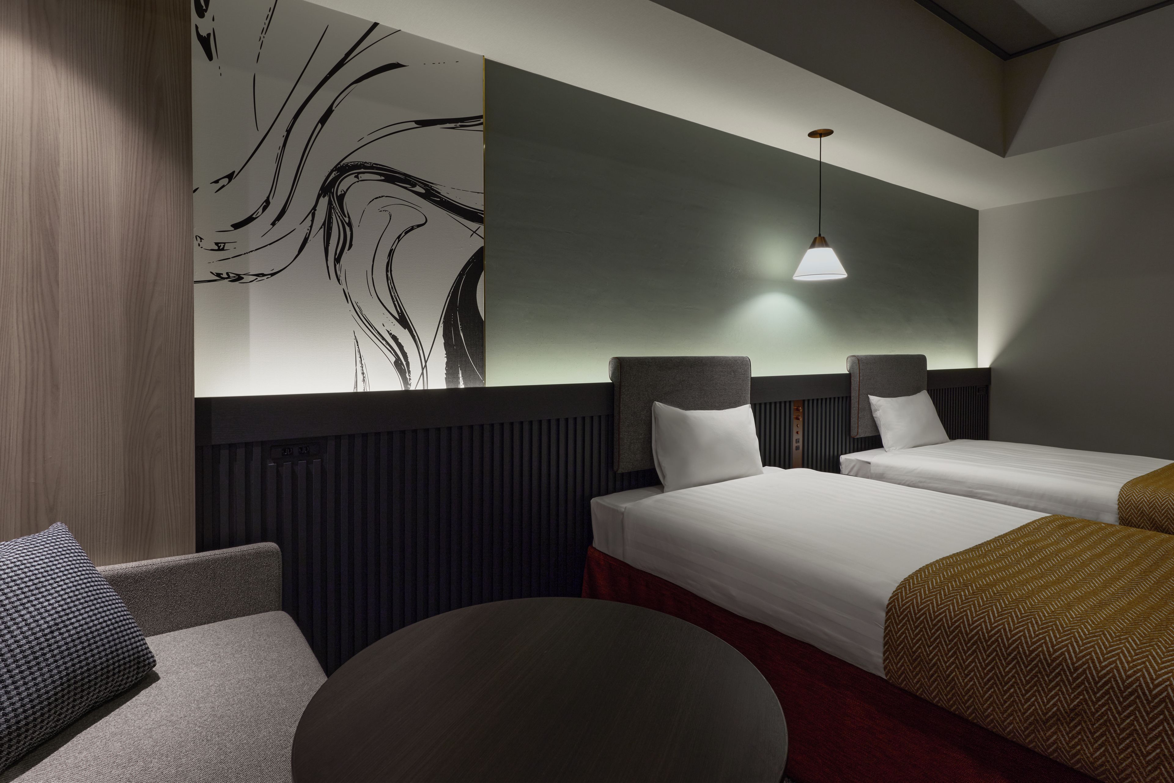 Quarto de hotel com duas camas, uma mesa redonda, um sofá e uma parede decorada com arte abstrata.