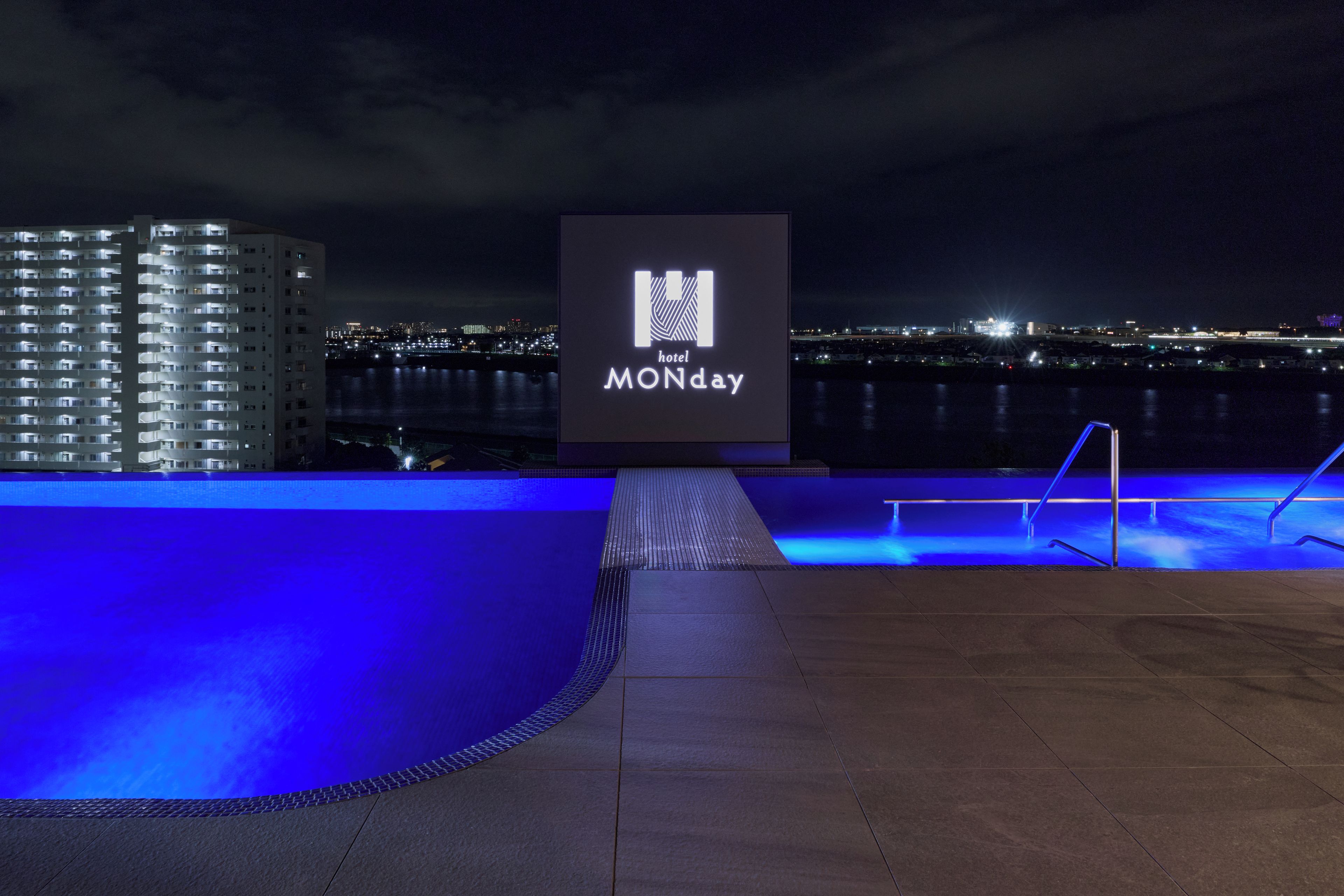 A nighttime view of a hotel with a pool area, a sign with the hotel's name, and a cityscape in the background.