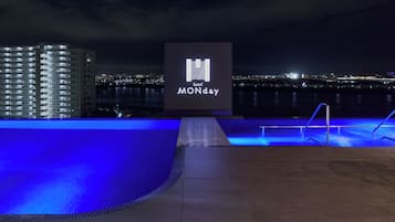 A nighttime view of a hotel with a pool area, a sign with the hotel's name, and a cityscape in the background.