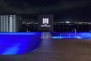 A nighttime view of a hotel with a pool area, a sign with the hotel's name, and a cityscape in the background.