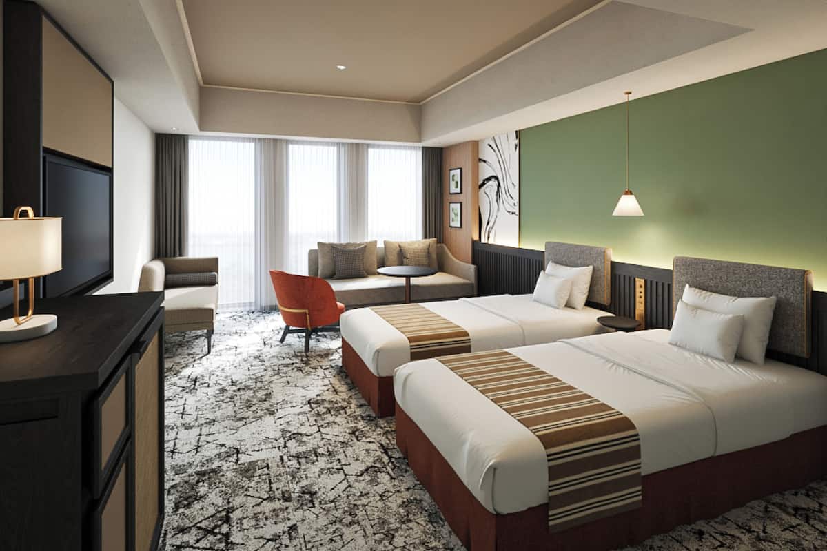 Habitación de hotel con dos camas, un escritorio, una silla, un televisor y un ventanal grande con cortinas.