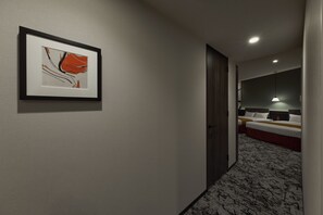 Ein Hotelkorridor mit einem gerahmten Kunstwerk an der Wand, einem Bett mit weißem Kopfteil und einem roten Akzent, sowie einem Teppichboden.