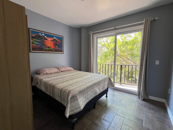 A bedroom with a bed, a painting, a balcony with a view of trees, and a window with curtains.