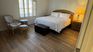 Ein Schlafzimmer mit einem Bett, einem Sessel, einem kleinen Tisch, einem Nachttisch mit Lampe, einem Deckenventilator und einem Fenster mit Jalousien.
