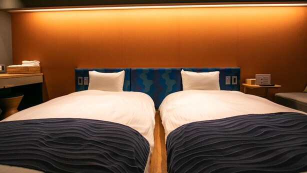 A hotel room with two beds, a headboard with a blue pattern, a bedside table with a lamp, and a small table with a device. - Minn Nijo-jo (Kyoto)