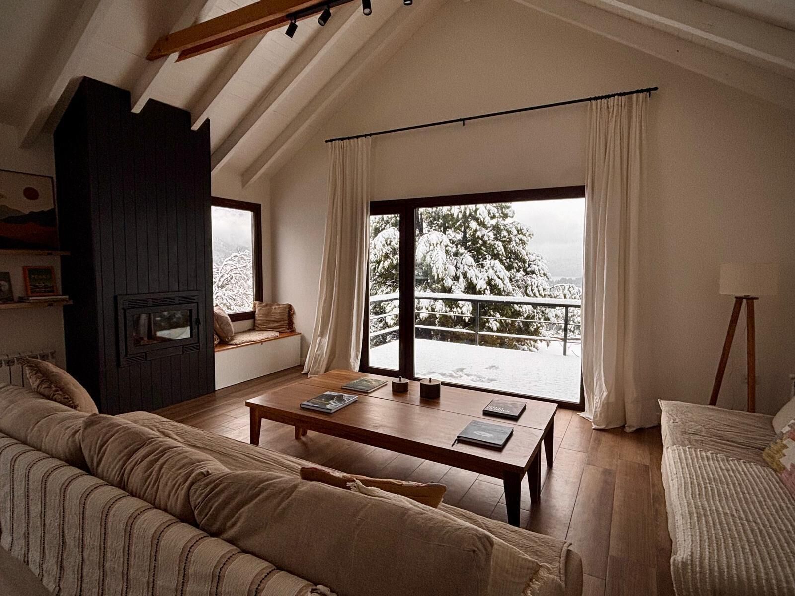 A cozy living room with a sofa, a coffee table, and a fireplace. There is a large window with a view of snow-covered trees.