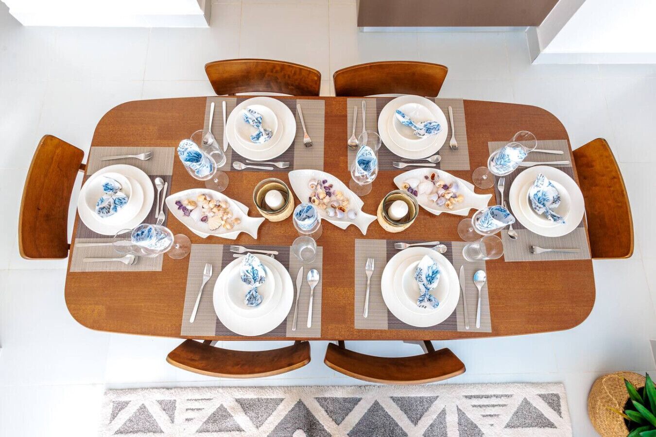 Mesa de comedor preparada para seis personas, con platos blancos y servilletas azules, rodeada de sillas de madera.