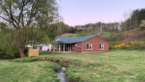 A single-story brick house with a sloped roof, a small porch, and a grassy yard with a stream. - Private 16-Acre Escape w/ Creek • Peaceful & Quiet • Fire Pit, Hiking & Lake (Hampton)