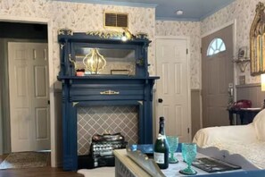 A room with a dark blue fireplace, a bed, a table with a bottle, glasses, and a book. - Crows Nest (Savannah)