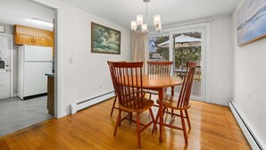 Uma área de refeições com mesa e cadeiras de madeira, luminária no teto e vista para um pátio externo através de uma porta de vidro.