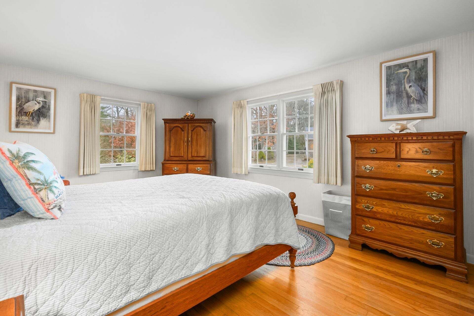 Une chambre à coucher comprenant un lit, une commode en bois, deux fenêtres avec des rideaux et des œuvres d’art murales.