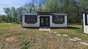 Eine kleine, eingeschossige Holzhütte mit Veranda, Fenstern mit schwarzen Rahmen und einem Metalldach.