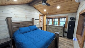 Ein Schlafzimmer mit Holzboden, einem Bett mit blauer Bettwäsche, einem Nachttisch, einer Kommode, einem Ventilator und Blick ins Freie durch ein Fenster.