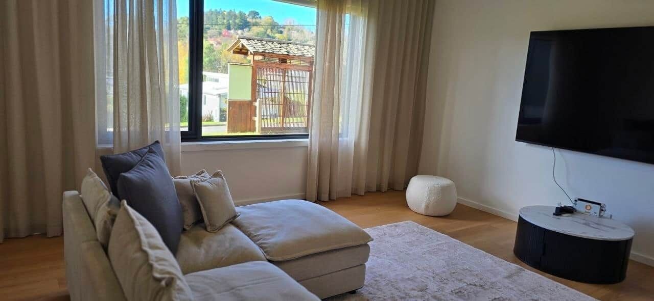 Una sala de estar con sofá, un reposapiés redondo, televisor de pantalla plana y una ventana con cortinas.
