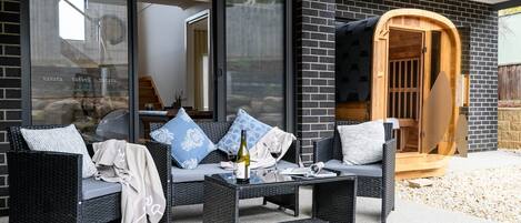 Un espacio al aire libre con mobiliario de mimbre, una mesa con una botella y vasos, y una puerta de sauna de madera.