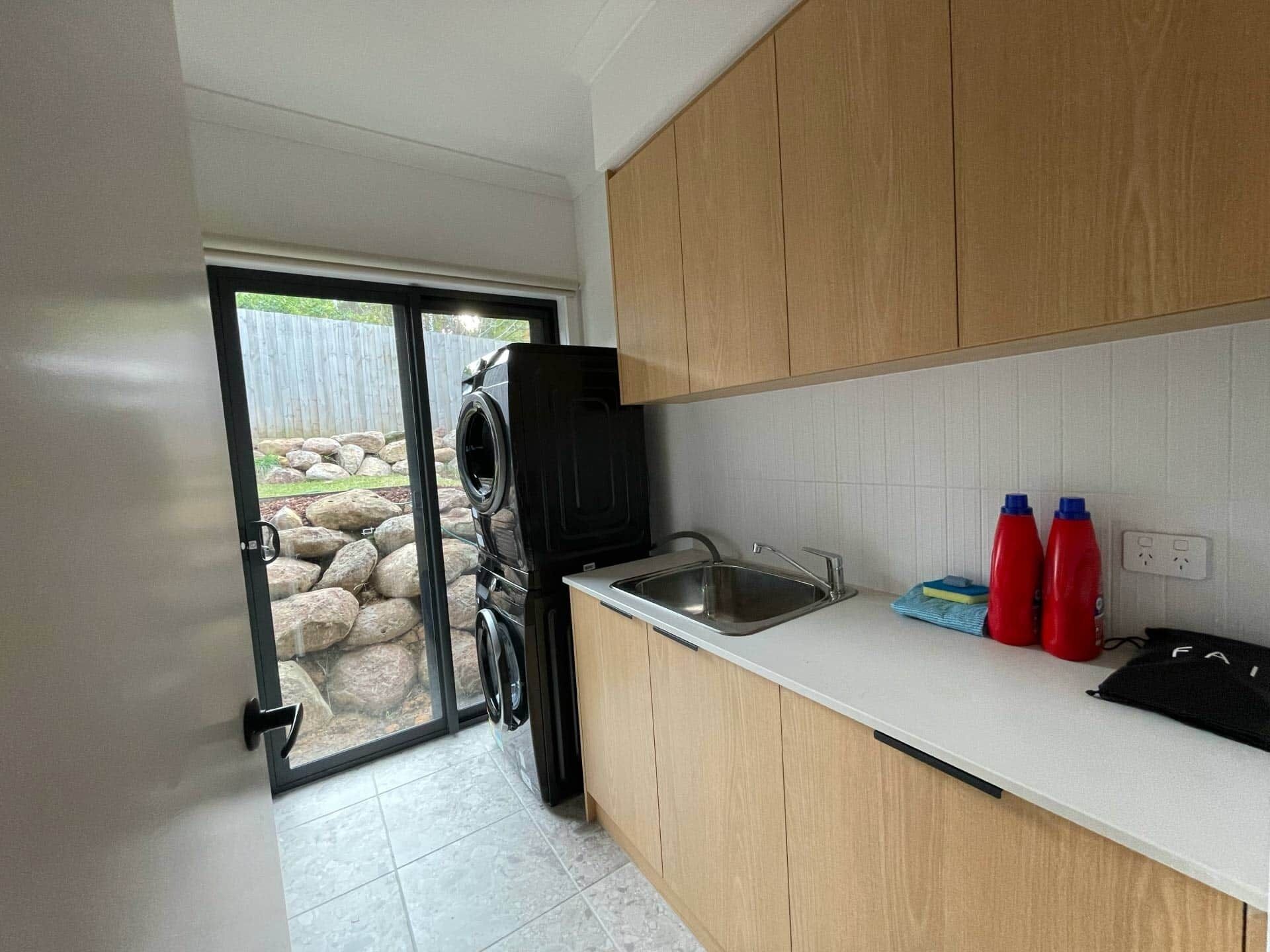 Una cocina con armarios de madera, un lavamanos de acero inoxidable y un portón de vidrio que da a un espacio exterior con rocas y una valla.