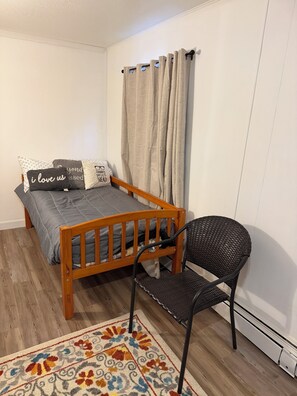 A small, cozy bedroom with a wooden bed, a black chair, a patterned rug, and a window with curtains. - Charming 2-bedroom apartment in Wasilla perfect for your getaway (Wasilla)
