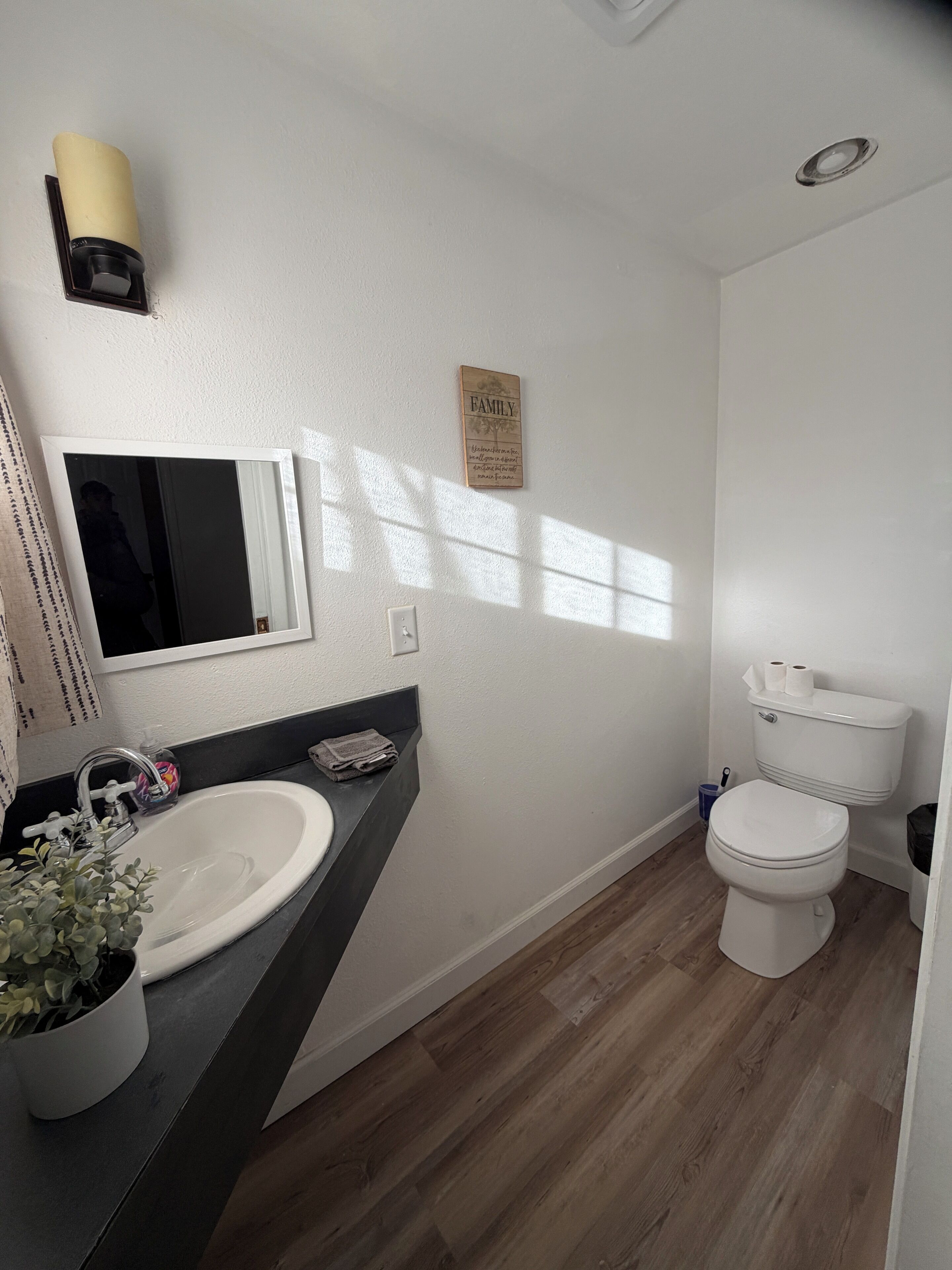 Ein Badezimmer mit weißem Waschbecken, Spiegel und Toilette. Eine Wandleuchte und ein Schild mit der Aufschrift 'FAMILY'.