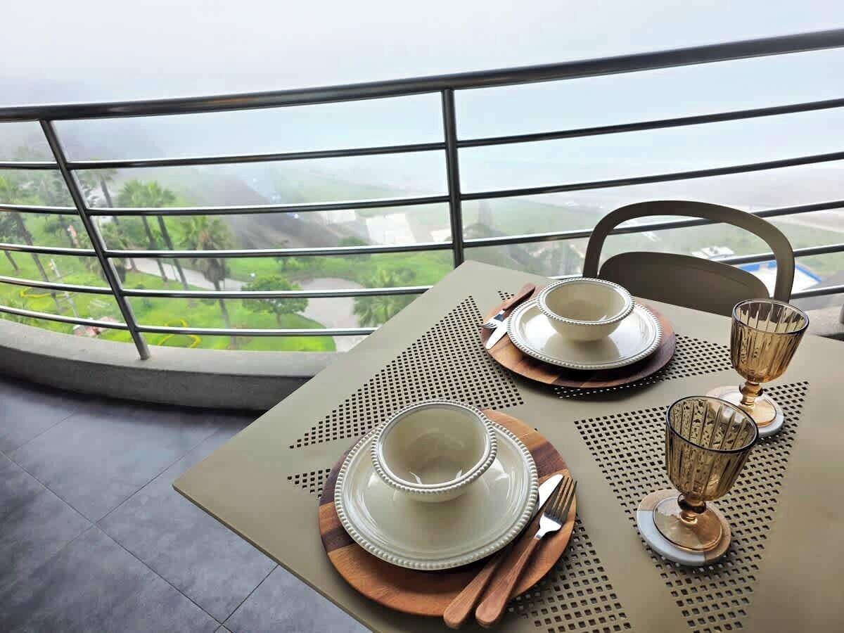 A table set with plates, bowls, and glasses, with a view of greenery and buildings through a window.