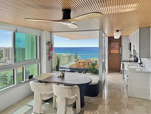 A modern kitchen with a view of the ocean, a dining area with a table and chairs, and a balcony with plants. - Sea to Sky Corner Penthouse Ocean and Diamond Head Views (Honolulu)