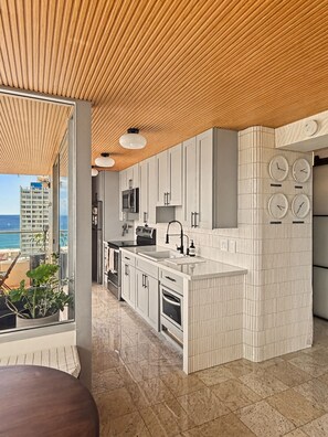 A modern kitchen with white cabinetry, a central island, a view of the ocean, and a wooden ceiling. - Sea to Sky Corner Penthouse Ocean and Diamond Head Views (Honolulu)