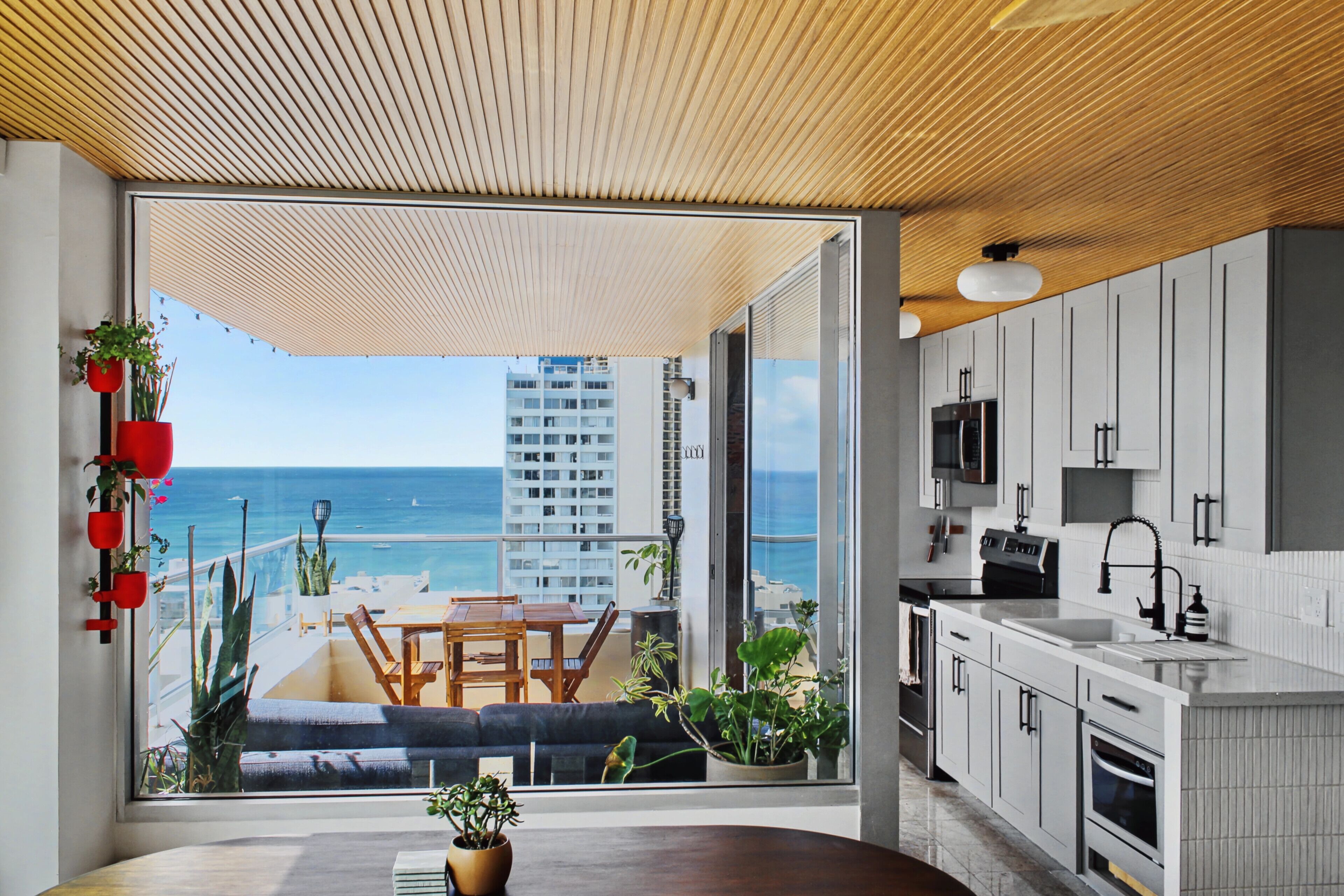 A modern kitchen with a view of the ocean, a dining area with a table and chairs, and a balcony with plants and a sofa.