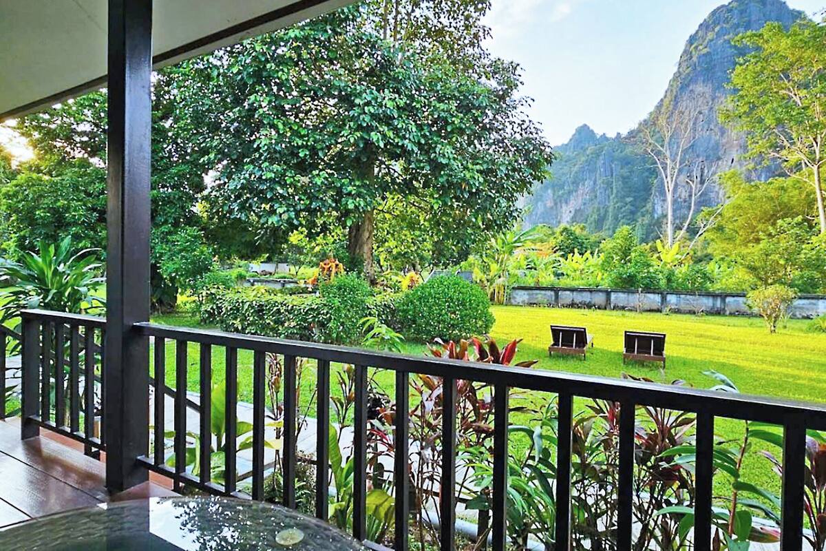 Un balcon donnant sur un jardin luxuriant, un petit étang et un paysage montagneux.