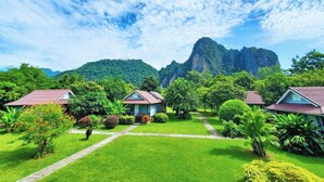 Un jardín exuberante con un sendero que lleva a un edificio, rodeado de follaje y una montaña al fondo.