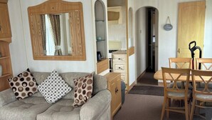 Ein Wohnzimmer mit einer Couch, gemusterten Kissen, einem Holzkabinett, einem Esstisch mit Stühlen und einer Küchenzeile im Hintergrund.