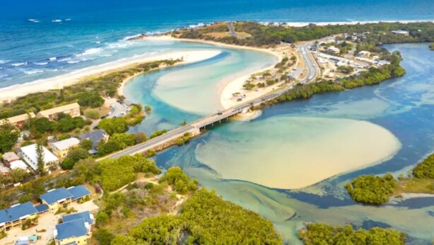 An aerial view of a coastal area with a beach, clear waters, and a bridge connecting to a peninsula with buildings and greenery.
