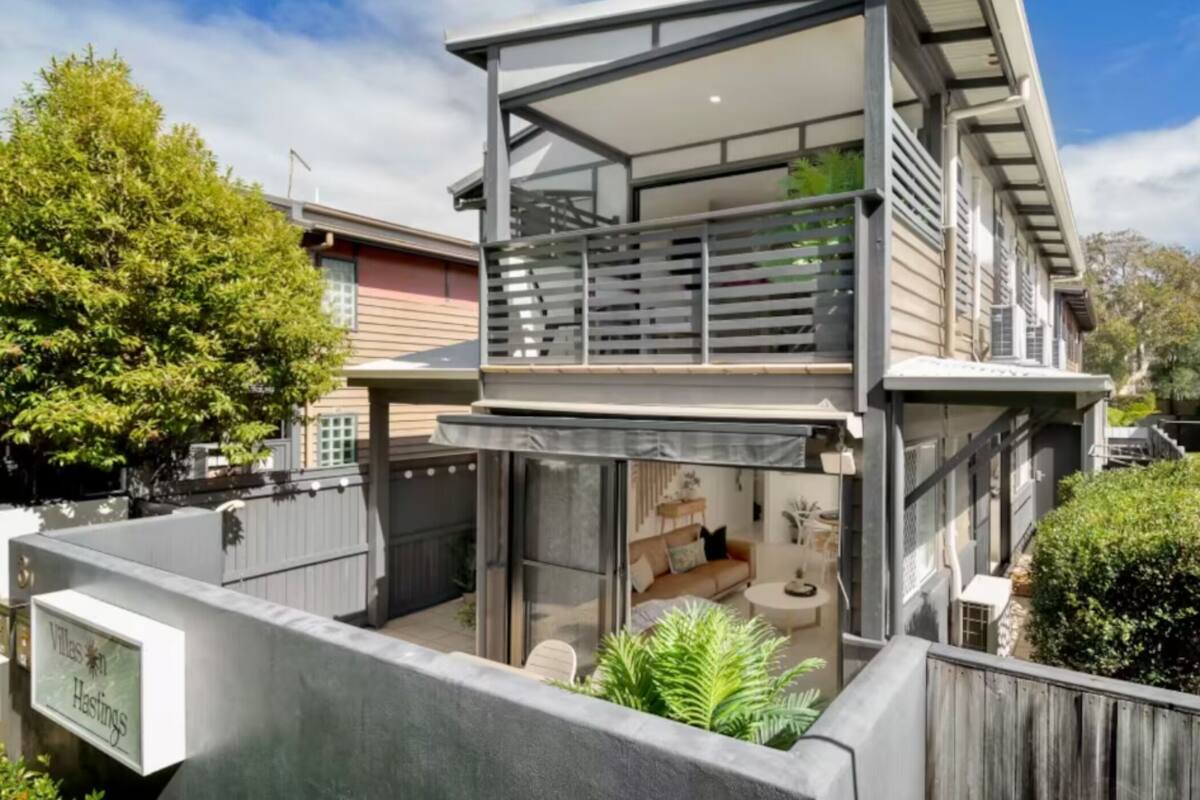 A modern two-story house with a balcony, a small patio, and a visible living area with a sofa and table.