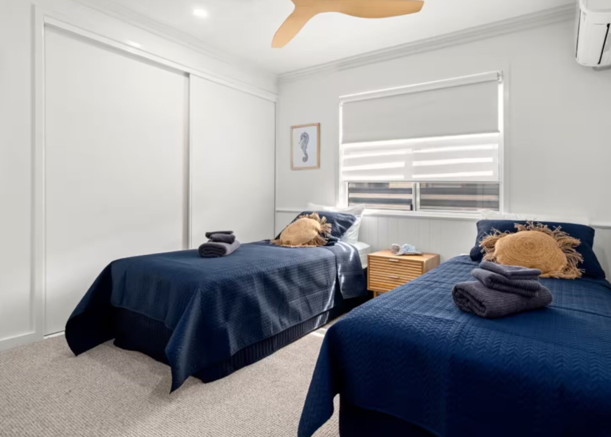 Une chambre avec deux lits, une fenêtre avec des stores, une table de chevet et un ventilateur de plafond.