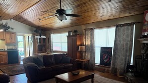 Ein Wohnzimmer mit braunem Ledersofa, einem Holztisch und einem Deckenventilator. Durch einen offenen Durchgang ist eine Küche zu sehen.