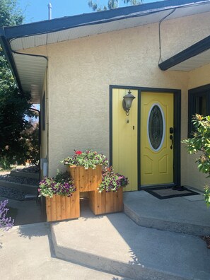 A building with a yellow door, a wooden planter with flowers, and a concrete staircase. - Summerland Bliss (Summerland)