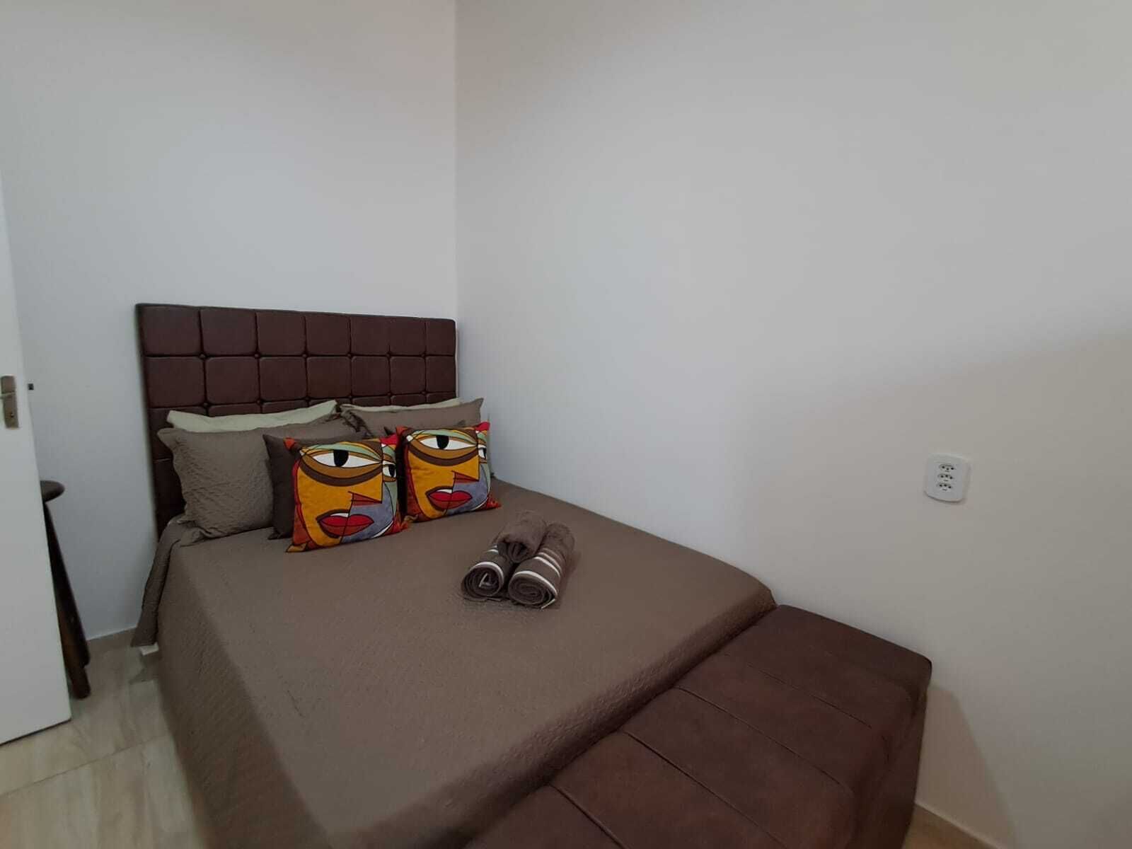 Un dormitorio con cabecera marrón, cama con colchón marrón, dos almohadas coloridas y un par de pantuflas sobre la cama.