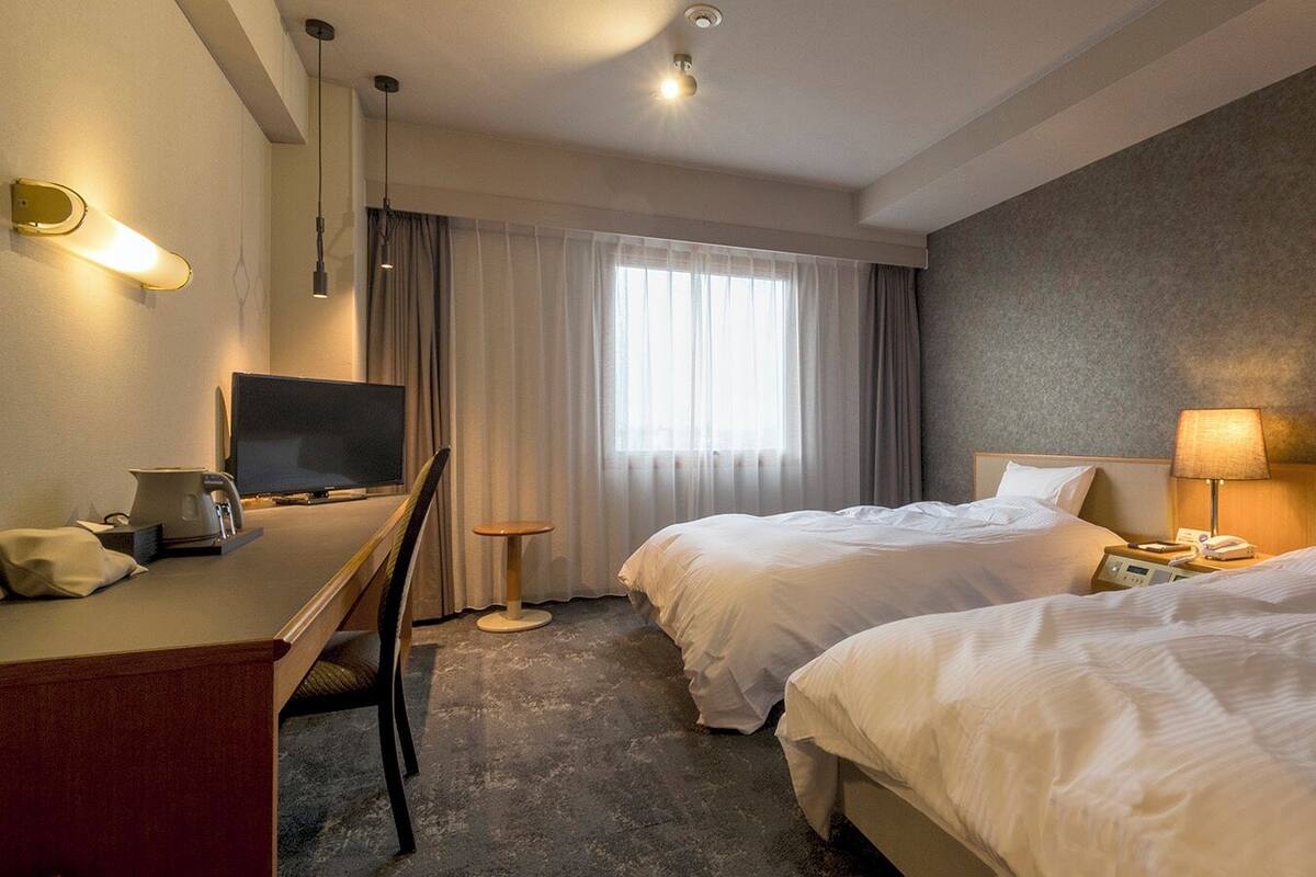 A hotel room with two beds, a desk with a computer, a chair, a lamp, and a TV.