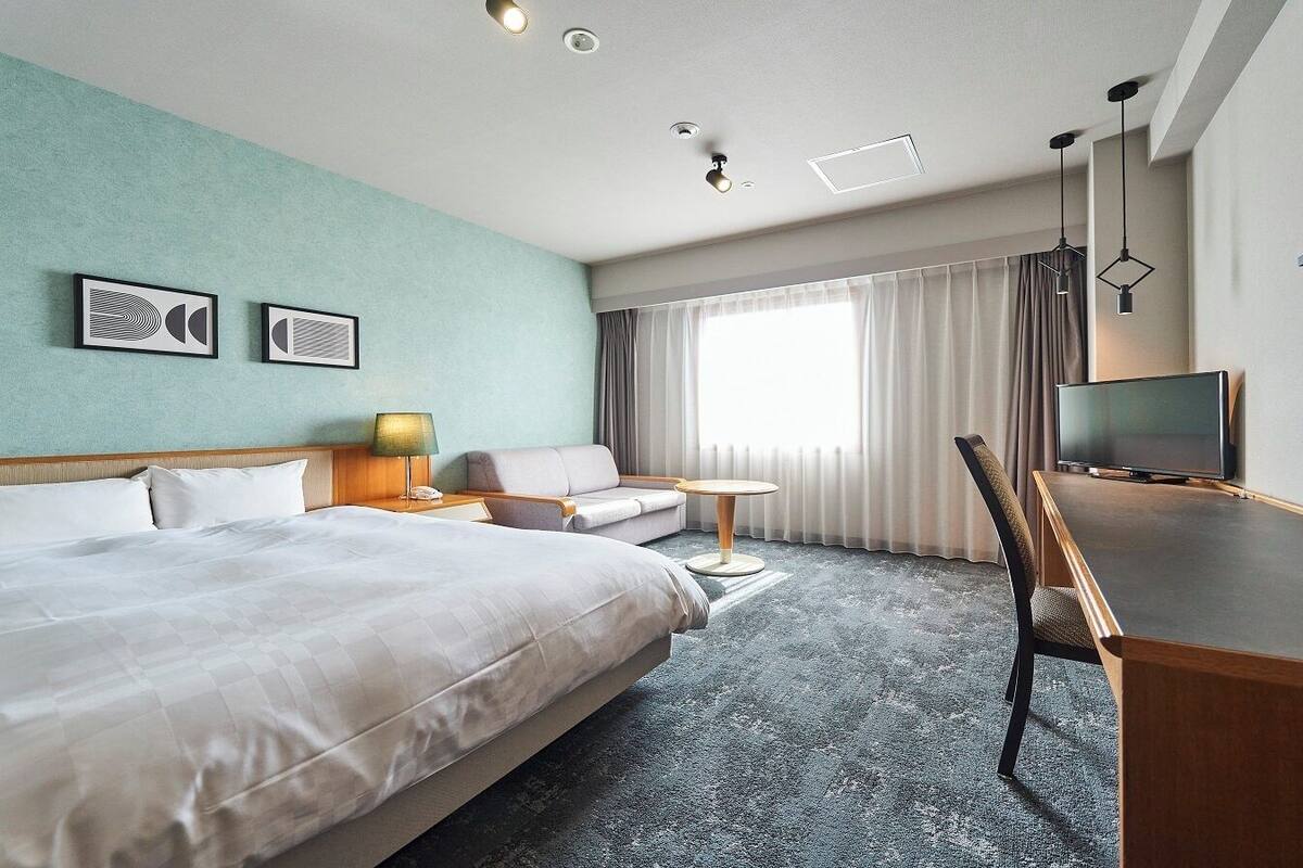 A hotel room with a large bed, a desk with a television, a chair, a small table, and a lamp.