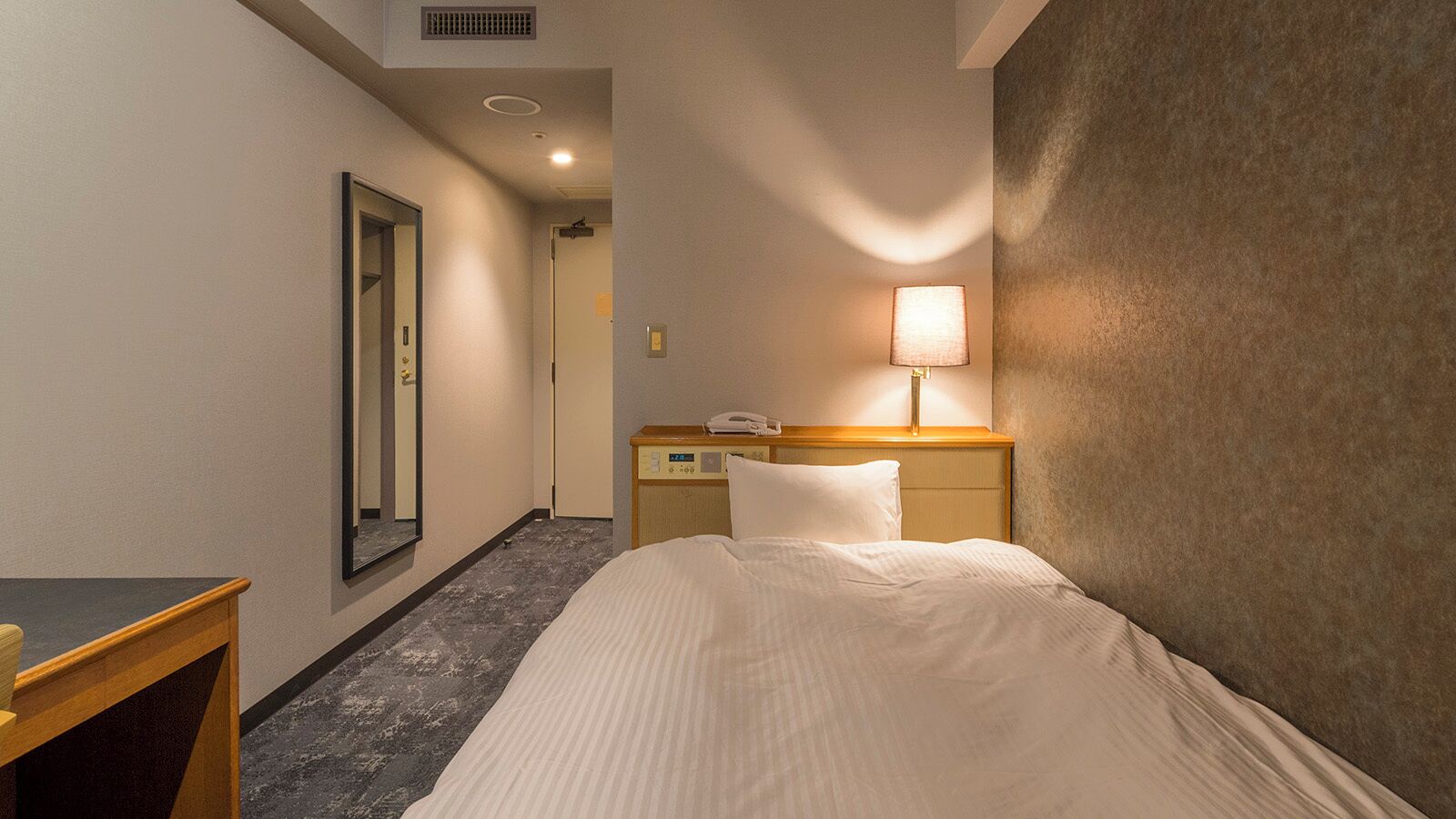 Una habitación de hotel con una cama, una mesita de noche, una lámpara, un espejo y una puerta que da a otra habitación.