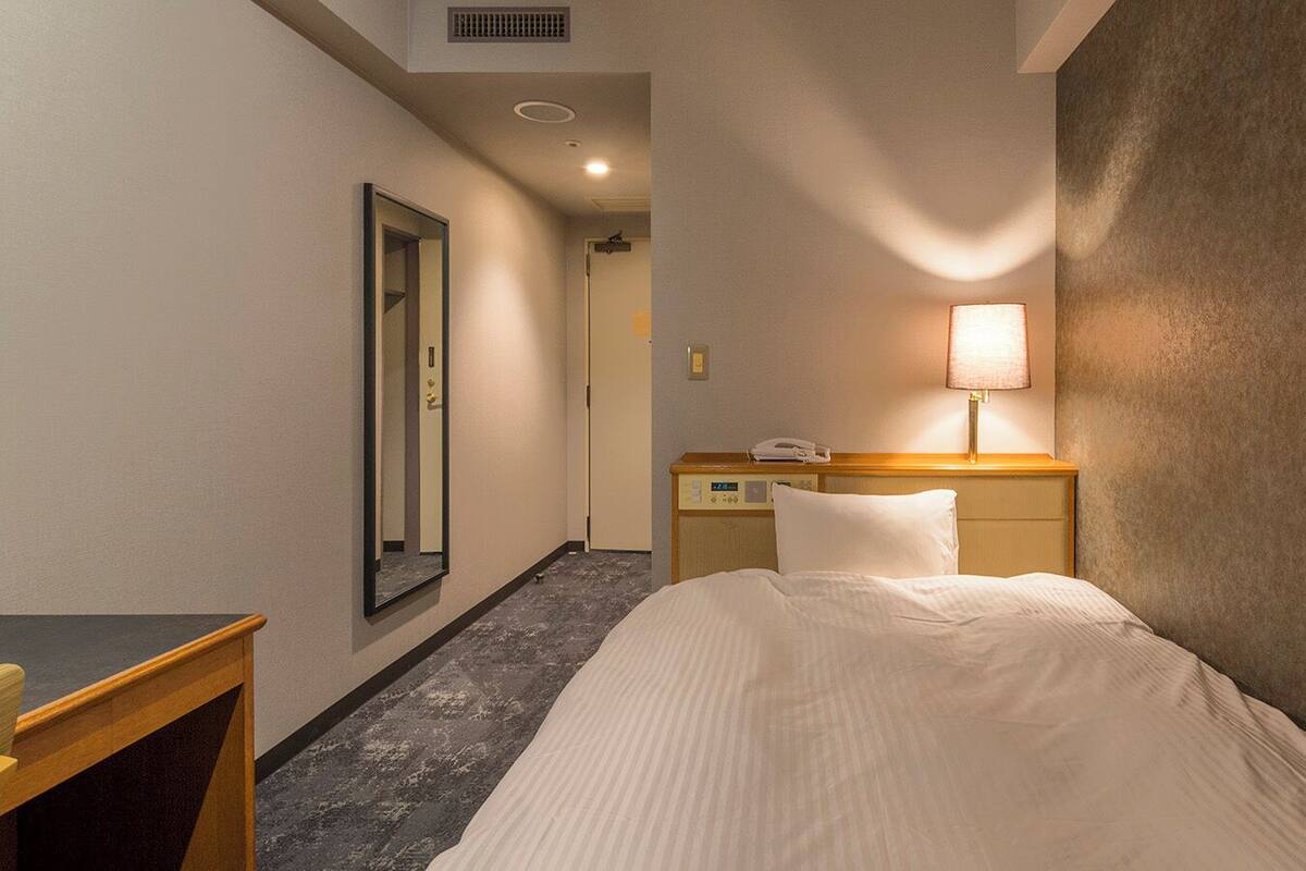 A hotel room with a bed, a bedside table, a lamp, a mirror, and a door leading to another room.