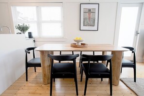 Une cuisine moderne avec une table à manger en bois, des chaises noires, un saladier rempli de fruits et une œuvre d’art encadrée accrochée au mur.