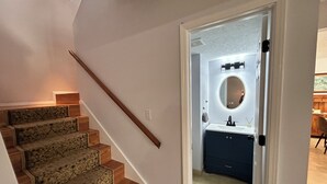 Un escalier recouvert d’une moquette à motifs mène à une salle de bain dotée d’un miroir rond et d’un meuble sombre.
