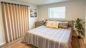 Een slaapkamer met een bed, een nachtkastje, een kamerplant en een raam met jaloezieën.